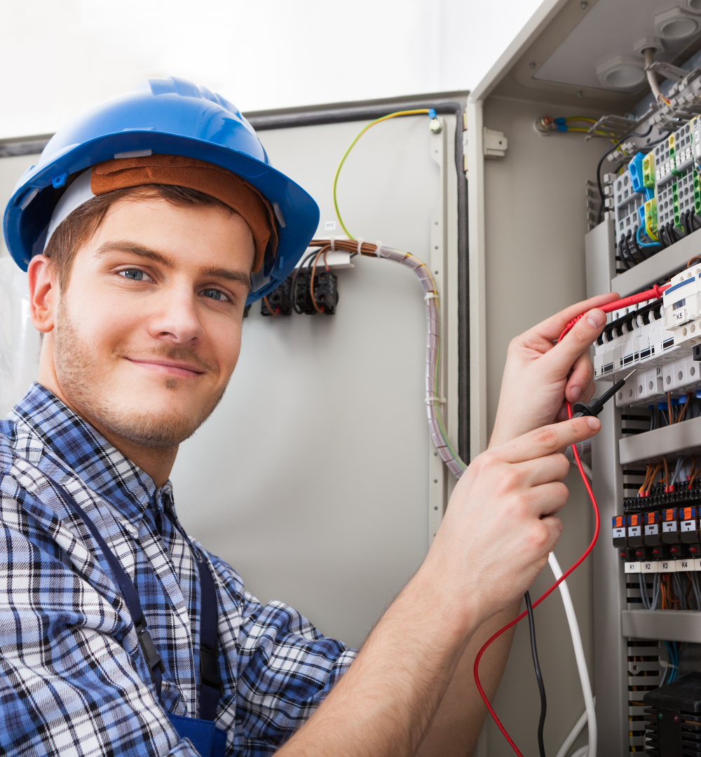 A man in a hard hat and plaid shirt works on an electrical panel, focused on his task in a construction setting.
